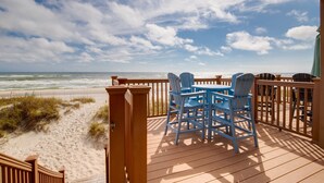Une terrasse en bois avec des fauteuils Adirondack bleus, donnant sur une plage de sable et des vagues.