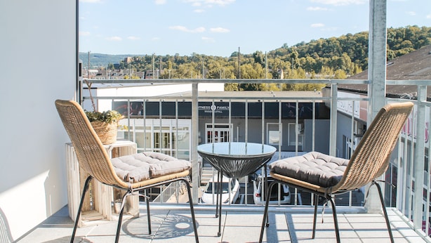 Un balcon avec des fauteuils en osier et une table en verre, d’où l’on peut admirer un immeuble et un paysage vallonné.