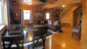 Interior de una cabaña de troncos con comedor, un conjunto de sofás y una escalera.