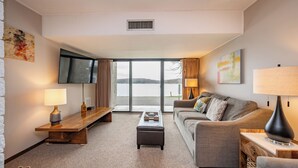 A modern living room with a large window, a flat-screen TV, a sofa, a coffee table, and two lamps. - Will O' The Wisp: 101B One Bedroom/One Bath (Oakland)