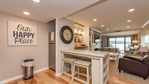 Uma sala de estar moderna com um sofá, um espaço de refeições com mesa e cadeiras, um relógio de parede grande e uma placa que diz 'Our Happy Place'.