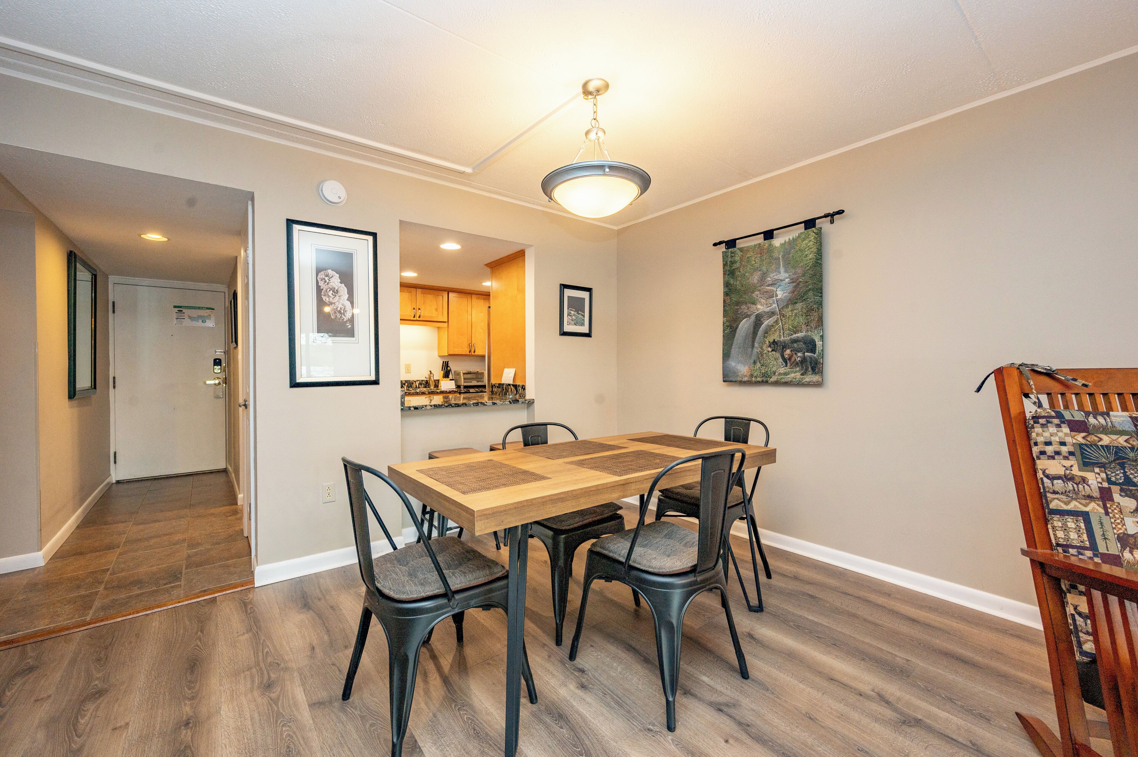A dining area with a wooden table and chairs, a kitchen in the background, and a hallway leading to another room.