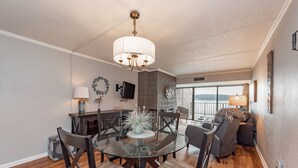Een moderne woonkamer met een glazen eettafel, donker houten stoelen, een kroonluchter en uitzicht op het omliggende landschap.