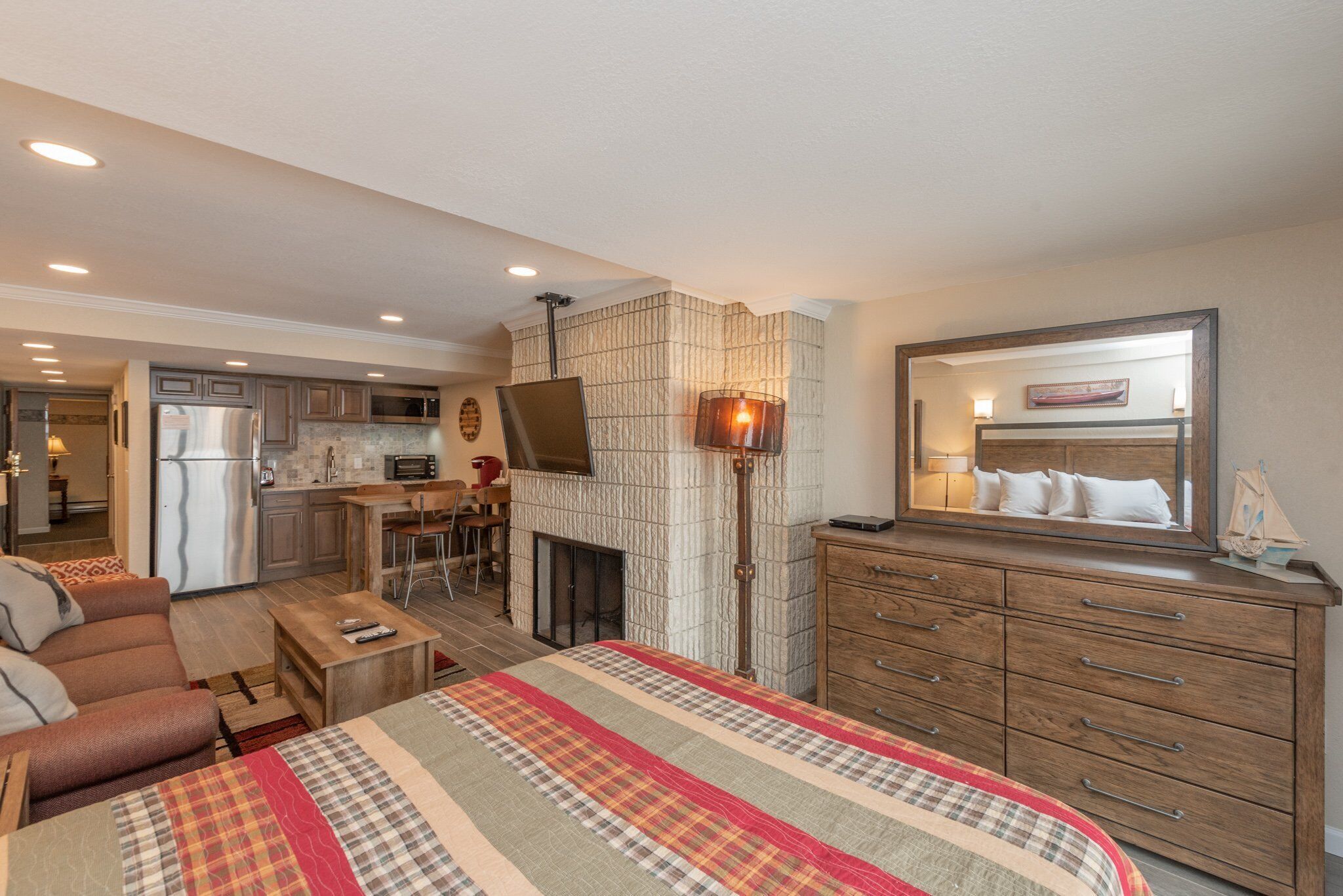 A bedroom with a large bed, a dresser, a nightstand, a fireplace, and a kitchen in the background.