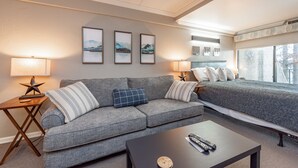Une chambre d’hôtel moderne avec un canapé gris, un lit recouvert d’une couvre-lit à motifs gris, une petite table avec une télécommande et des tableaux encadrés accrochés au mur.