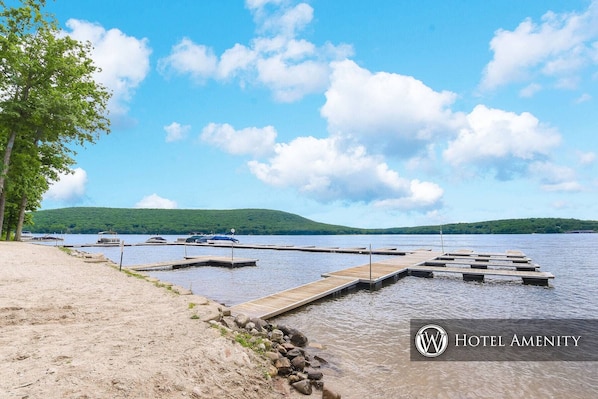 Guests can enjoy a serene lakeside setting with a dock extending into the water, surrounded by a sandy beach area and rolling hills in the distance.