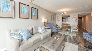 A living room with a sofa, armchairs, and a coffee table. There is a dining area with a table and chairs. The room has a chandelier and artwork on the walls. - Will O' The Wisp: 301D Two Bedroom/Two Bath (Oakland)
