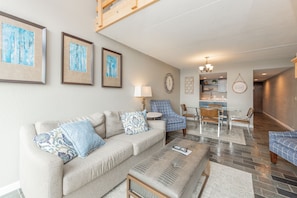 A living room with a sofa, armchairs, and a coffee table. There is a dining area with a table and chairs. The room has a chandelier and artwork on the walls. - Will O' The Wisp: 301D Two Bedroom/Two Bath (Oakland)