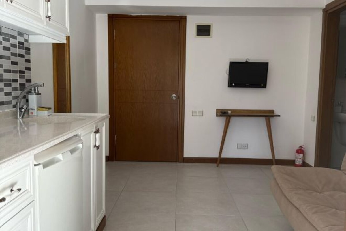 Une cuisine avec des meubles blancs, un lave-vaisselle et un crédence carrelée. On y trouve une porte en bois, un téléviseur fixé au mur et une petite table avec une chaise.