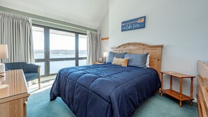 Habitación de hotel con una cama grande, cabecera de madera, vista al mar, escritorio con lámpara y una silla.