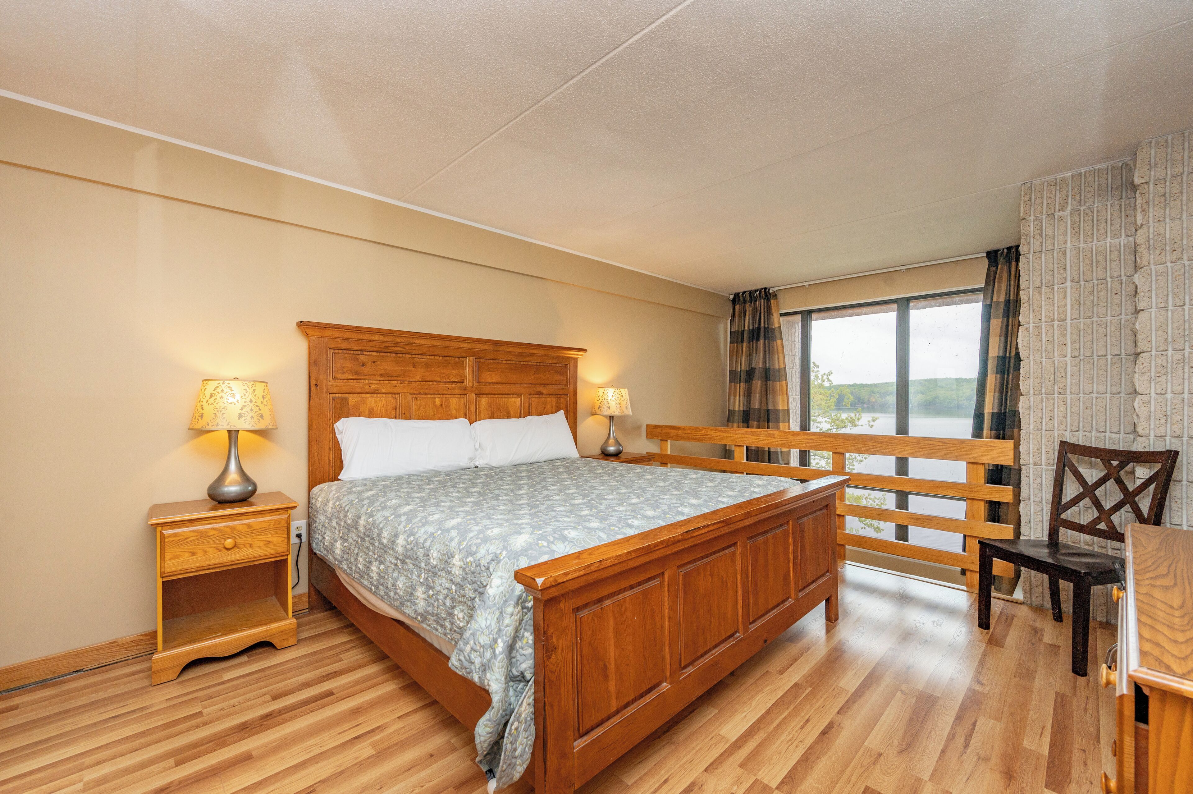 Un dormitorio con cama de madera, dos mesitas de noche con lámparas, un banco de madera y vistas al exterior a través de una ventana.