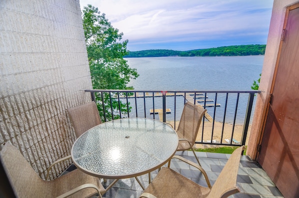 A balcony with a glass table and chairs, overlooking a lake with a dock and a sandy beach.