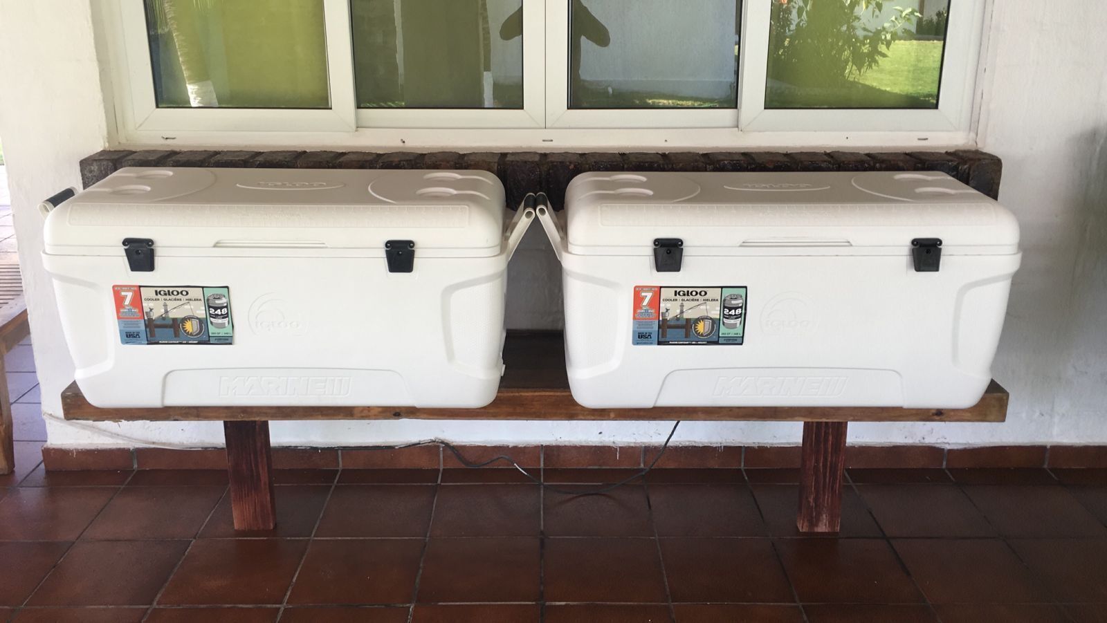 Zwei weiße, transportable Kühlboxen mit dem Schriftzug 'IGI100' auf einer Holzbank vor einer fensterdurchbrochenen Wand.