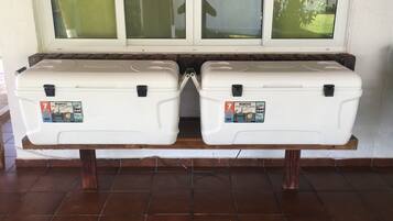 Zwei weiße, transportable Kühlboxen mit dem Schriftzug 'IGI100' auf einer Holzbank vor einer fensterdurchbrochenen Wand.
