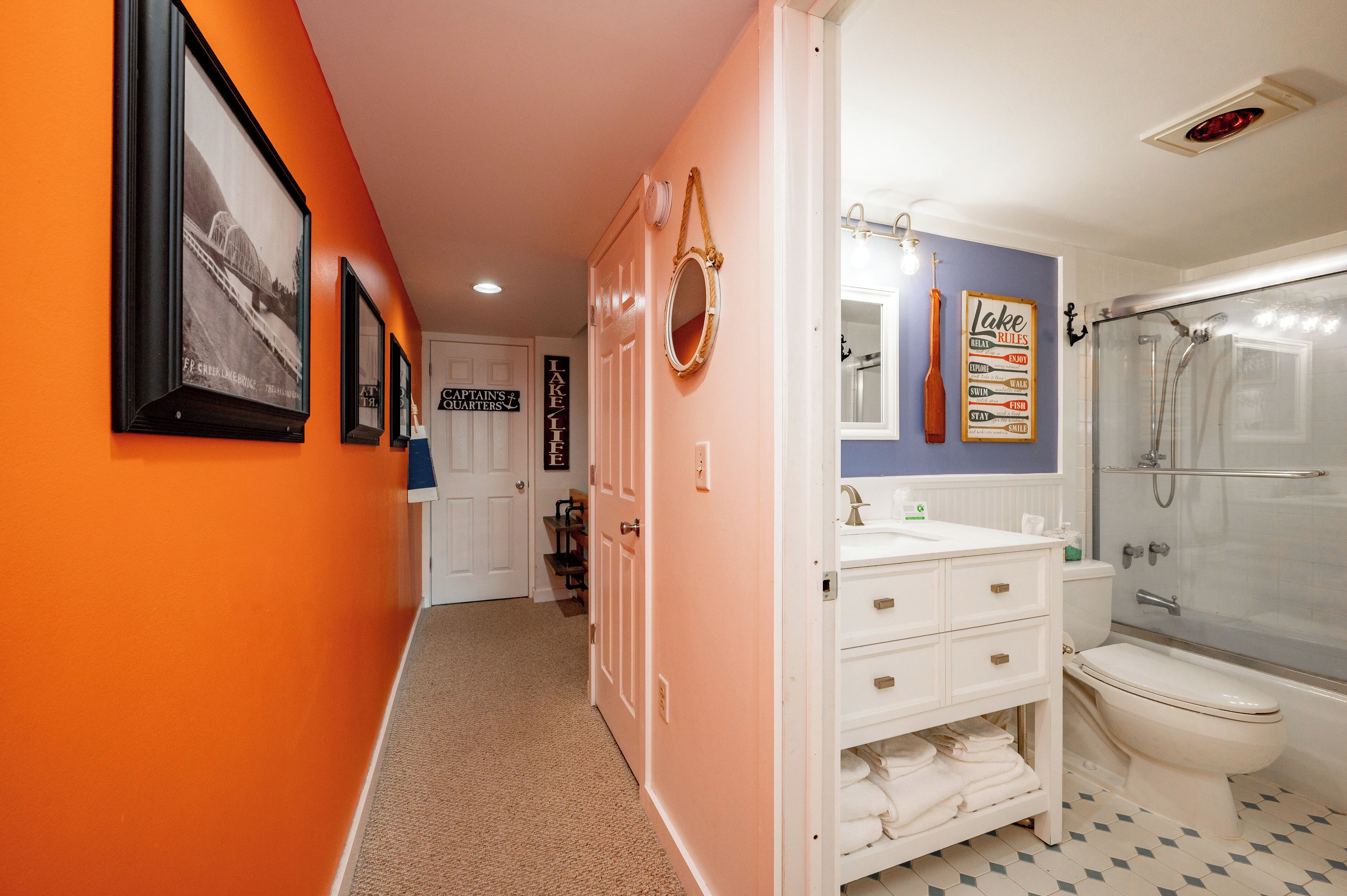 Een gang die leidt naar een badkamer met een witte wastafel, toilet en douche. Er hangen foto’s in lijsten aan de muur en een bord met de tekst “Captains' Quarters”.