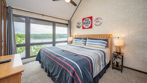 A bedroom with a large bed, a ceiling fan, a nightstand with a lamp, and a view of a lake through the window. - Will O' The Wisp: 708H Studio Loft/One Bath (Oakland)