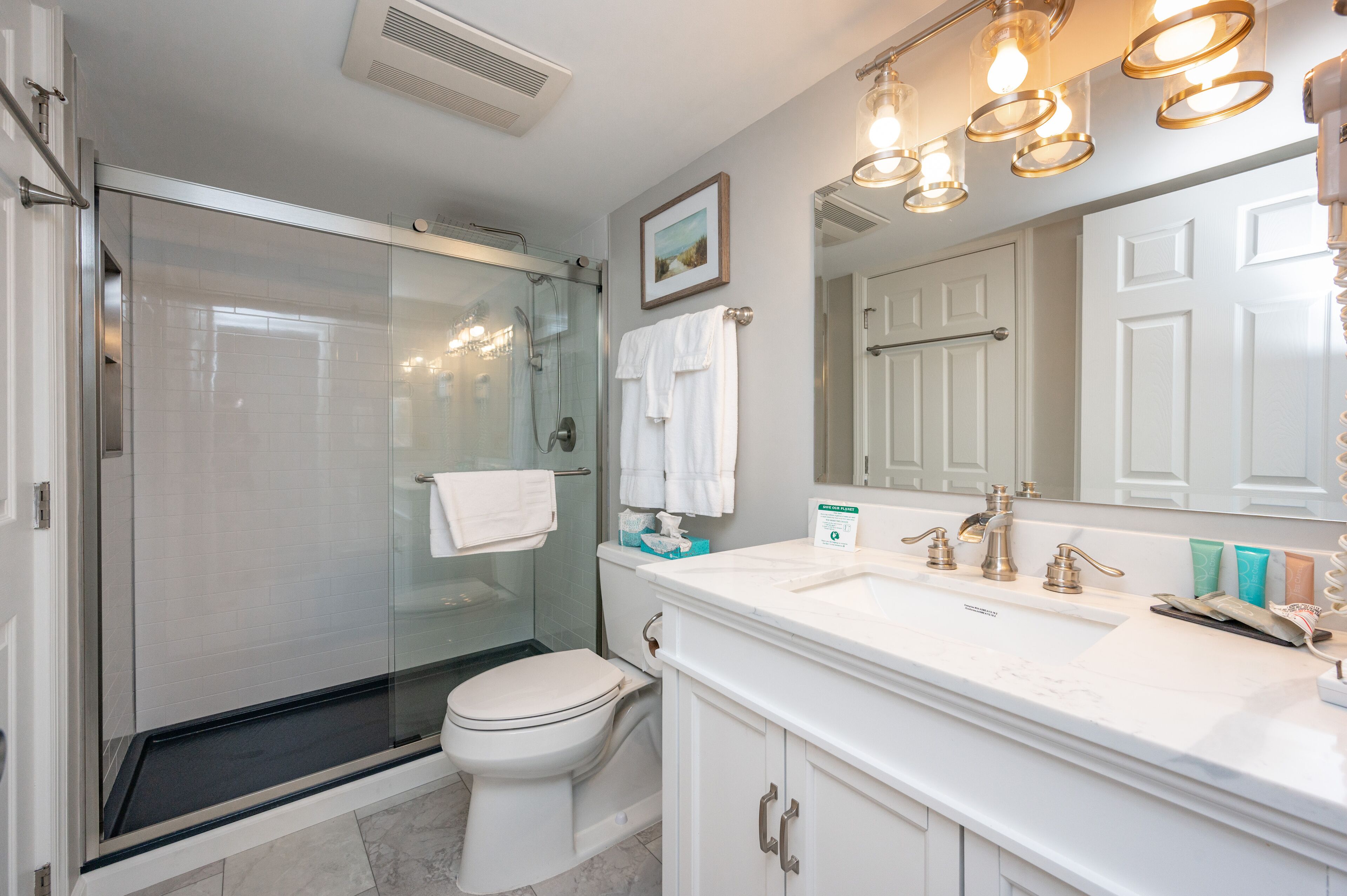 A modern bathroom with a glass-enclosed shower, a white toilet, and a vanity with a large mirror and multiple light fixtures.