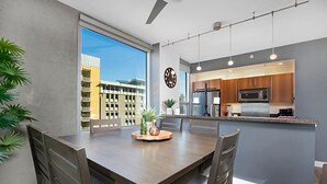 Eine moderne Küche mit einer zentralen Kochinsel, einem Essbereich mit Holztisch und Stühlen sowie Blick auf ein Gebäude durch ein großes Fenster.