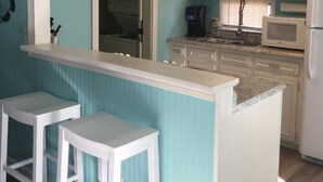 A kitchen with a teal wall, white cabinets, a white countertop, and two white bar stools. - Waterfront, Large deck, Pet friendly, Golf cart, Screened in porch. (Myrtle Beach)