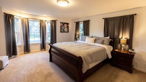 A bedroom with a large bed, dark wooden headboard, beige carpet, and a nightstand with a lamp. - Indoor Heated Pool & Hot Tub, Game room, EV Charger, Pets Welcome! (Bend)