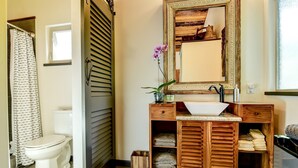 Une salle de bain avec un meuble-lavabo en bois, un lavabo blanc et un miroir. On y trouve également des toilettes, un rideau de douche et une plante en pot posée sur le meuble-lavabo.