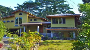 Une maison à deux étages, à la façade jaune, dotée d’un porche et d’un jardin bien entretenu où poussent différentes espèces de plantes et où s’étend une pelouse.