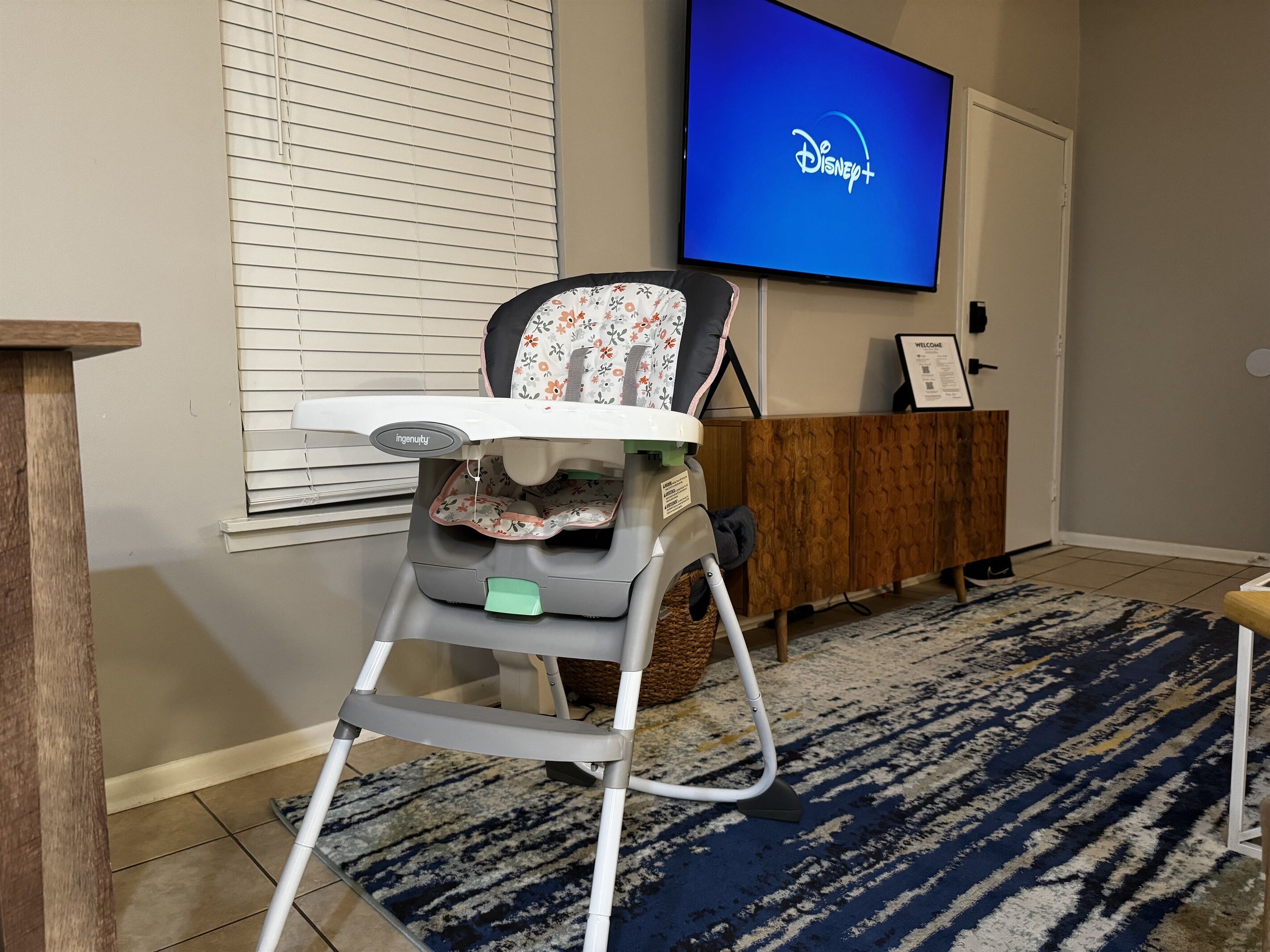 A baby high chair in a room with a television displaying the Disney+ logo, a wooden cabinet, and a patterned rug.