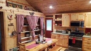 A cozy cabin-style kitchen with wooden cabinets, a wooden dining table with benches, a red and white checkered curtain, and a wall-mounted clock. - Couples Retreat, hot tub, private! (La Pine)