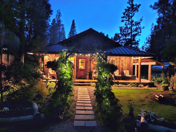A wooden house with a porch, surrounded by trees and a pathway leading to the entrance.