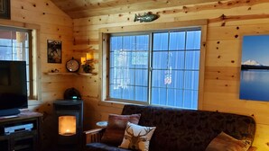 A cozy cabin interior with a wooden ceiling, a sofa with patterned cushions, a television, a fireplace, and a large window offering a view of a snowy mountain. - Couples Retreat, hot tub, private! (La Pine)