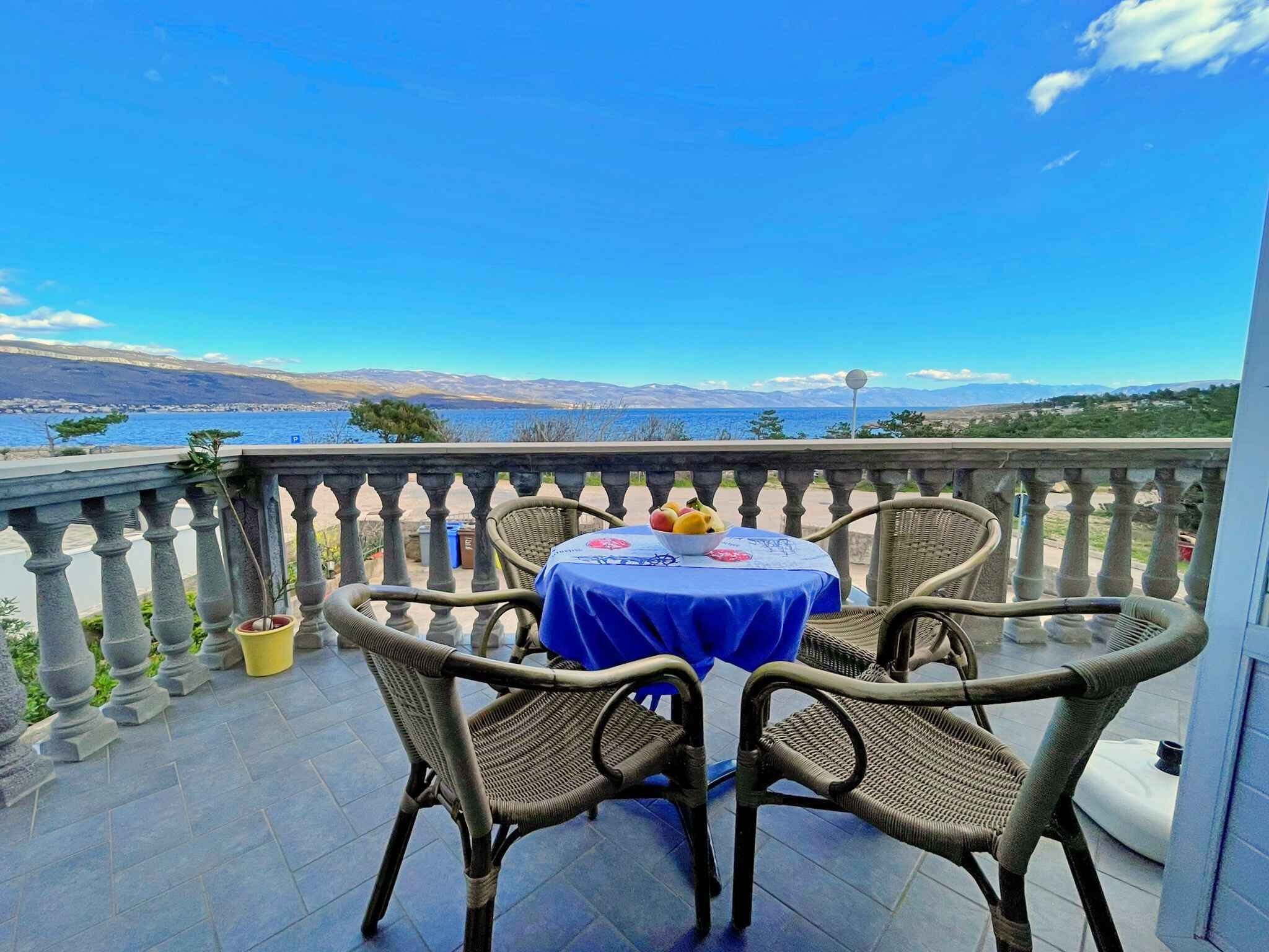 A balcony with wicker chairs and a table, overlooking a scenic view of the sea and distant mountains.