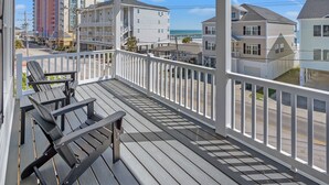 A covered porch with two Adirondack chairs and a picnic table, overlooking a beach and residential buildings. - Modern Luxury Home w/ Private Pool - Cherry Grove Beach (North Myrtle Beach)