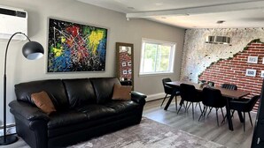 A living room with a black leather sofa, a coffee table, a dining table with chairs, a large abstract painting, and a brick wall. - Stylish 3BR Home Close to Campus 10min to Boston (Everett)