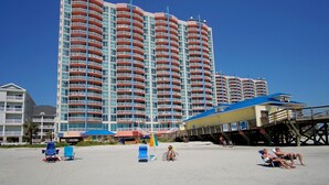 A high-rise hotel with multiple balconies, a beach area with sun loungers, and a pier structure.