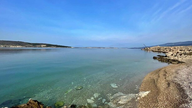 Vista despejada de un paisaje costero con mar tranquilo, costa rocosa y colinas a lo lejos.