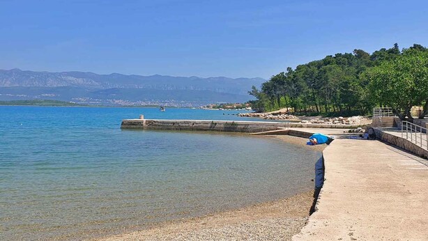 Un paysage côtier avec une mer bleue et limpide, une plage de galets et une jetée qui s’avance dans l’eau.