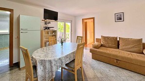 Un espace de vie compact comprenant une table à manger, des chaises, un canapé, un téléviseur à écran plat fixé au-dessus de la table, et une petite kitchenette équipée d’un réfrigérateur.