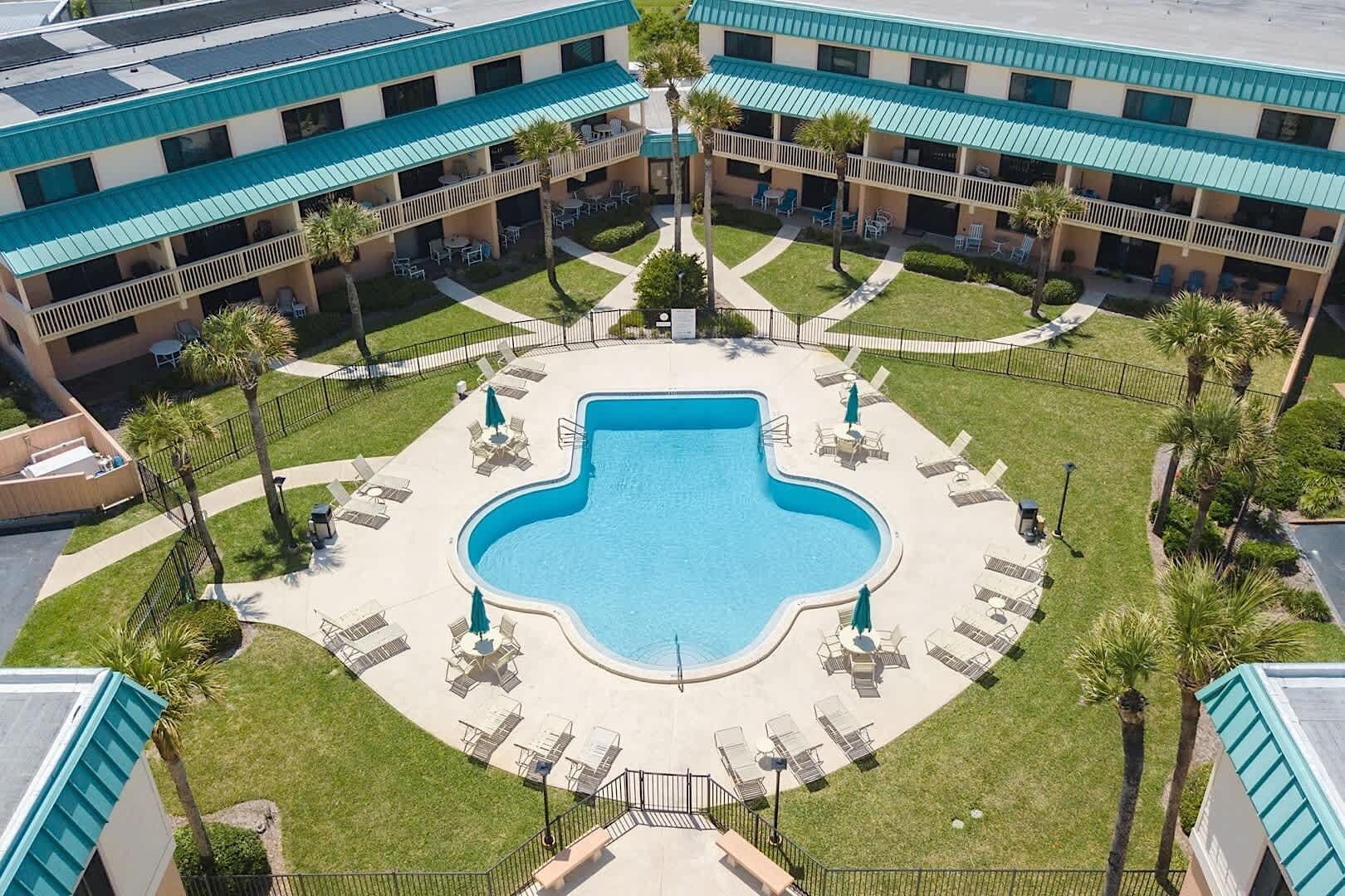 A hotel with a central swimming pool surrounded by lounge chairs and palm trees.