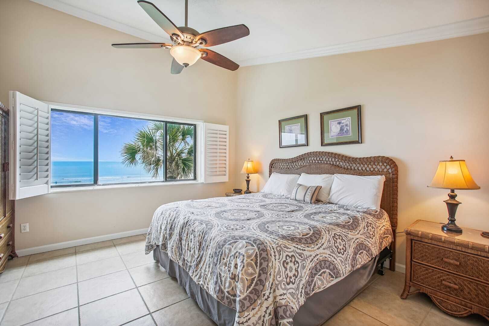 A bedroom with a large bed, a ceiling fan, a window with shutters, two bedside lamps, and a nightstand.