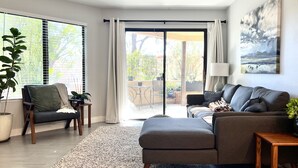 A modern living room with a grey sofa, a wooden coffee table, and a large window with blinds. - Contemporary Condo with Private Garden Patio and Pool Access (Tucson)