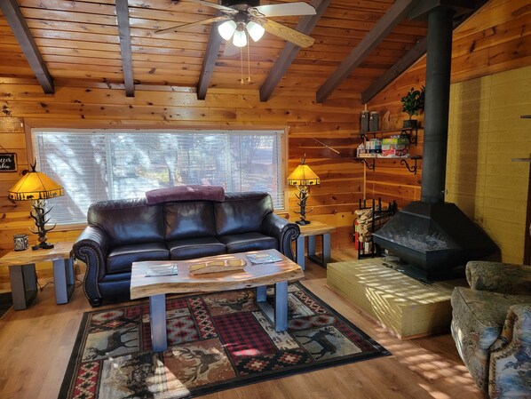 Ein gemütliches Blockhauss Innenleben mit Ledersofa, Holztisch, gemustertem Teppich, Deckenventilator und Kamin.