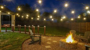 An outdoor patio area with a fire pit, Adirondack chairs, and a string of lights. - Big D Dallas Home : Hot Tub, Fire pit, large yard (Dallas)
