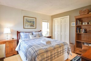 Quarto com cama, mesas de cabeceira, um aparador e uma estante de livros, além de um quadro na parede.