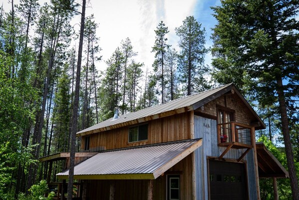 A cabin with a wooden exterior, a metal roof, and a balcony. It is surrounded by tall trees.