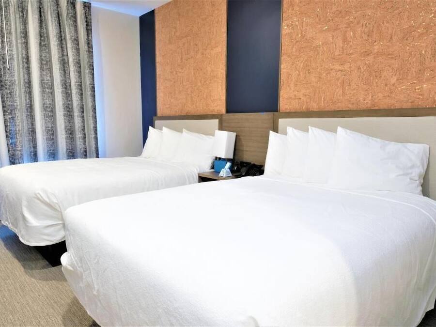 A hotel room with two beds, a nightstand, and a wall-mounted air conditioning unit.