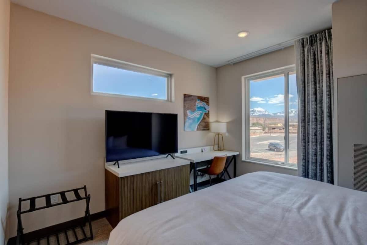 A hotel room with a bed, a desk with a chair, a television, and a window with a view of mountains.