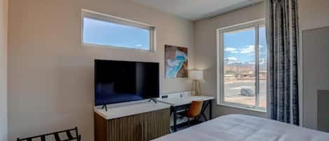 A hotel room with a bed, a desk with a chair, a television, and a window with a view of mountains.
