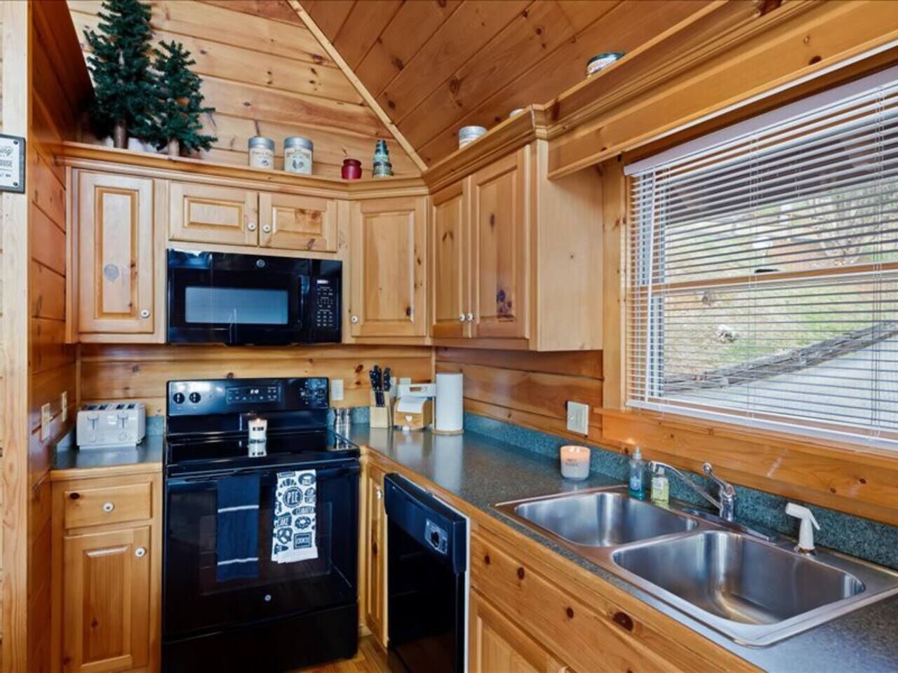 Eine kompakte Küche mit Holzschränken, einer Mikrowelle, einem Backofen, einem Spülbecken und einem Fenster mit Jalousien.