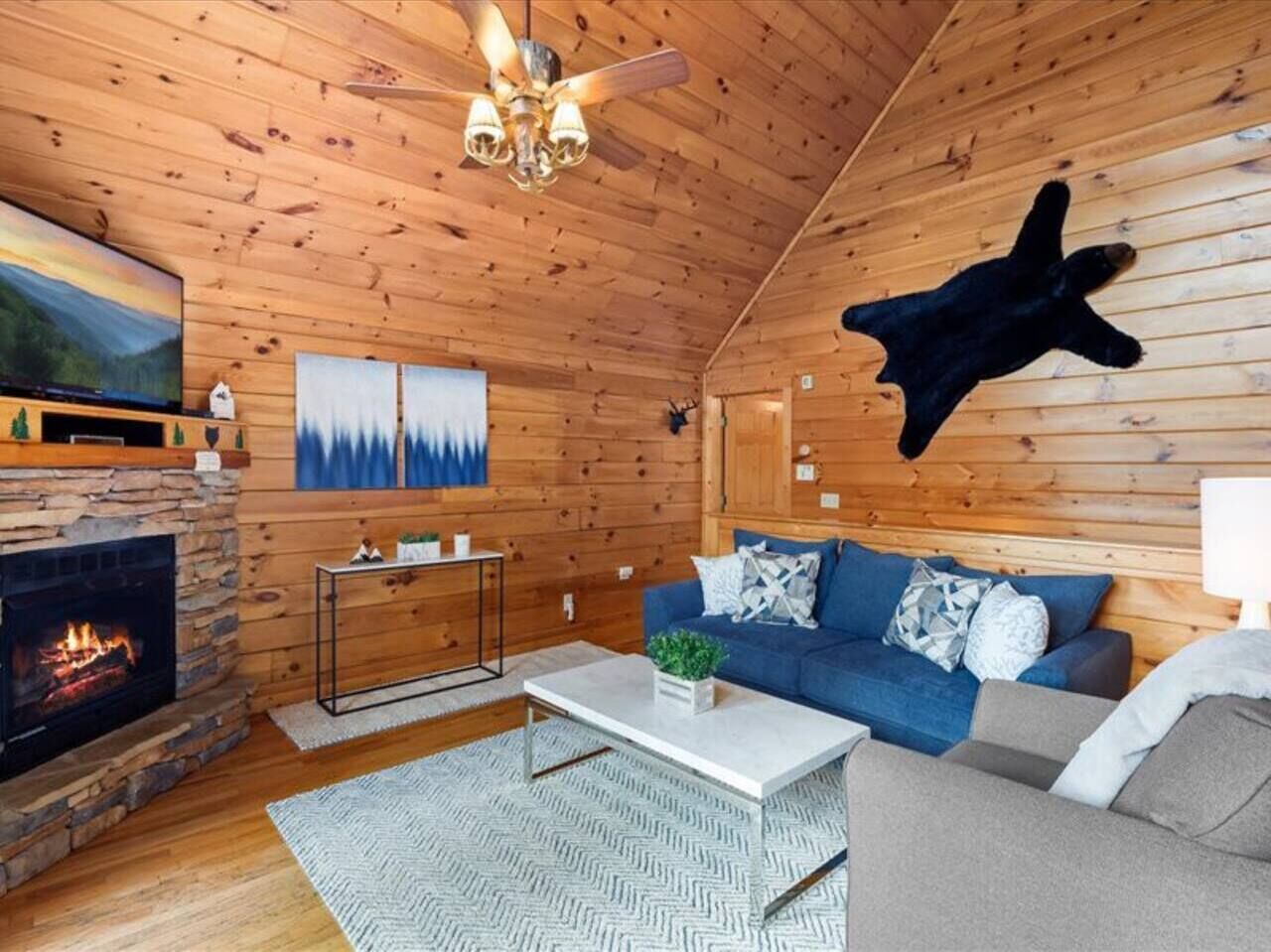 A cozy log cabin interior with a stone fireplace, a blue sofa, a white coffee table, and a ceiling fan.