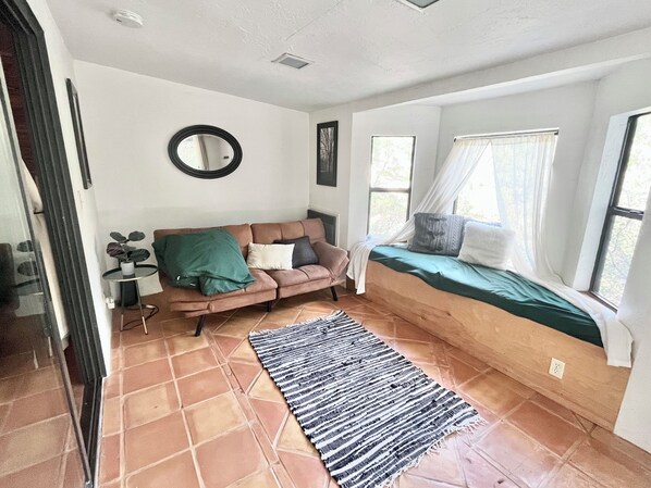 A cozy room with a sofa, a window with a bed, a mirror, and a small table with a plant.